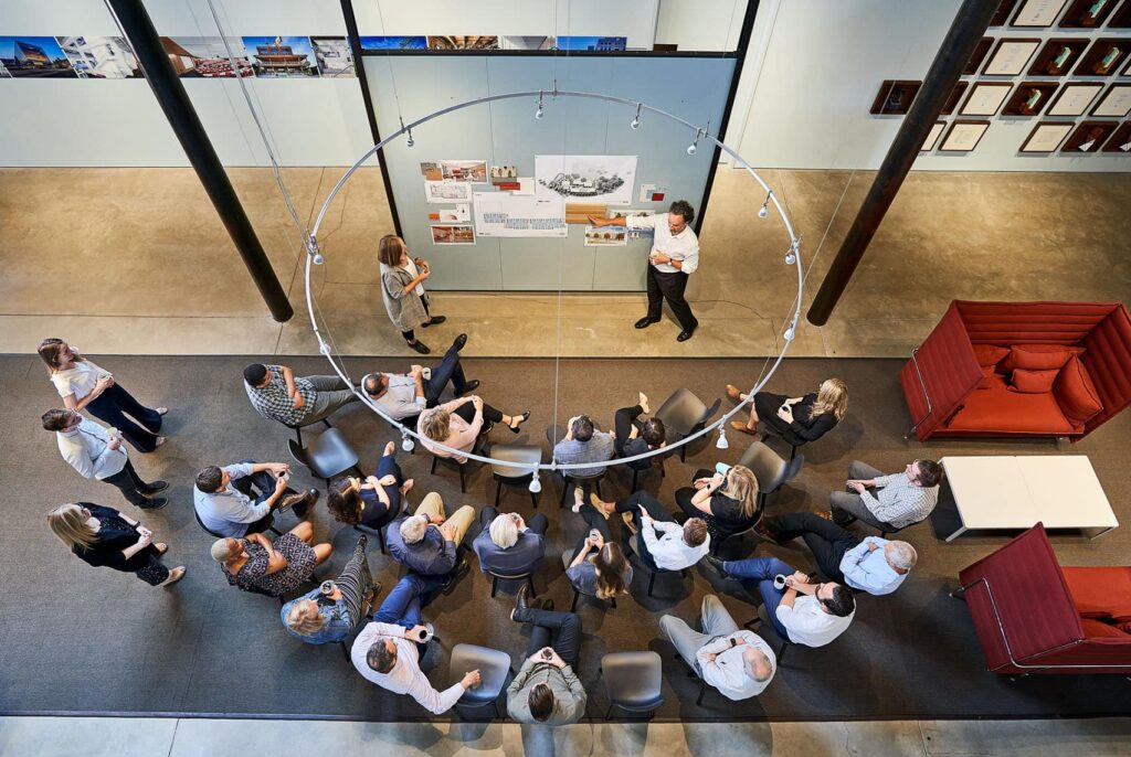 A group of employees seated around a pin-up board while others give a presentation.